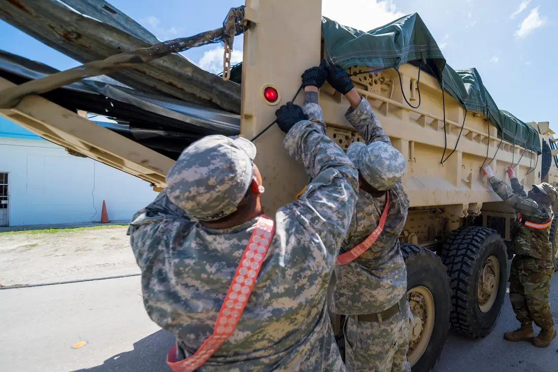 Soldiers pulling straps to secure a green tarp over a dump truck loaded with storm debris.