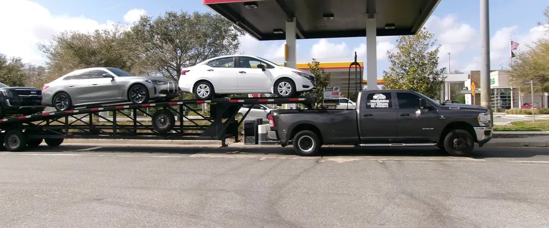 Dark Ram dually pickup pulling a gooseneck car hauler loaded with cars, stopped at a fuel station.