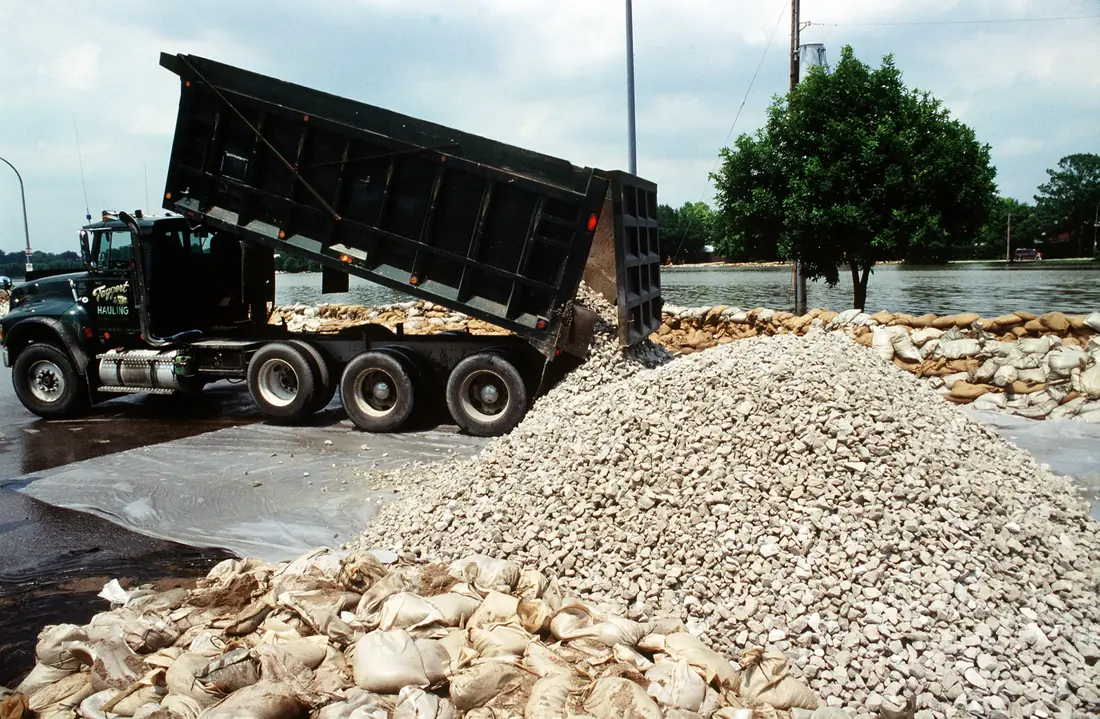 Dump truck with its bed raised, dumping crushed stone onto a pile beside a sandbag wall.