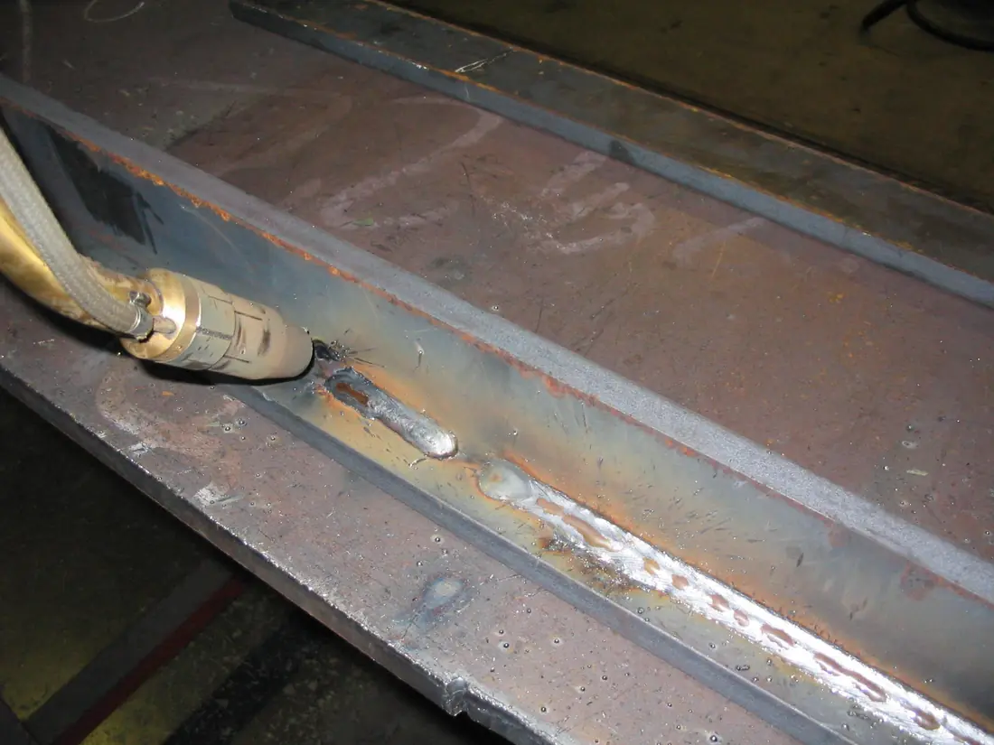 Welding torch next to a shiny fillet weld running along the inside corner of a steel channel.
