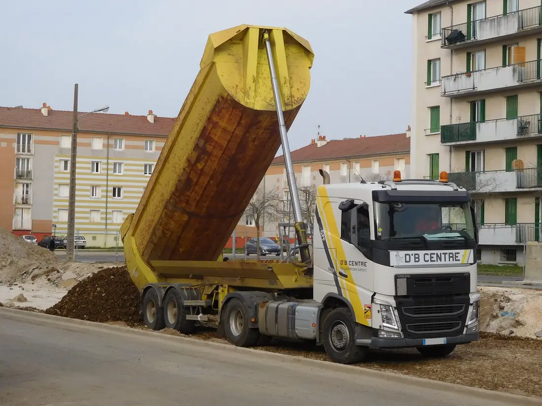 Tipper semi-trailer dumping dirt, its body raised by a long multi-stage cylinder mounted at the front of the body.