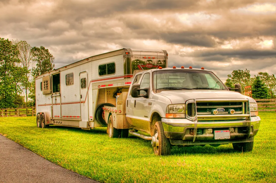 Silver Ford heavy-duty pickup with a flatbed, hitched to a tandem-axle gooseneck horse trailer parked on grass.