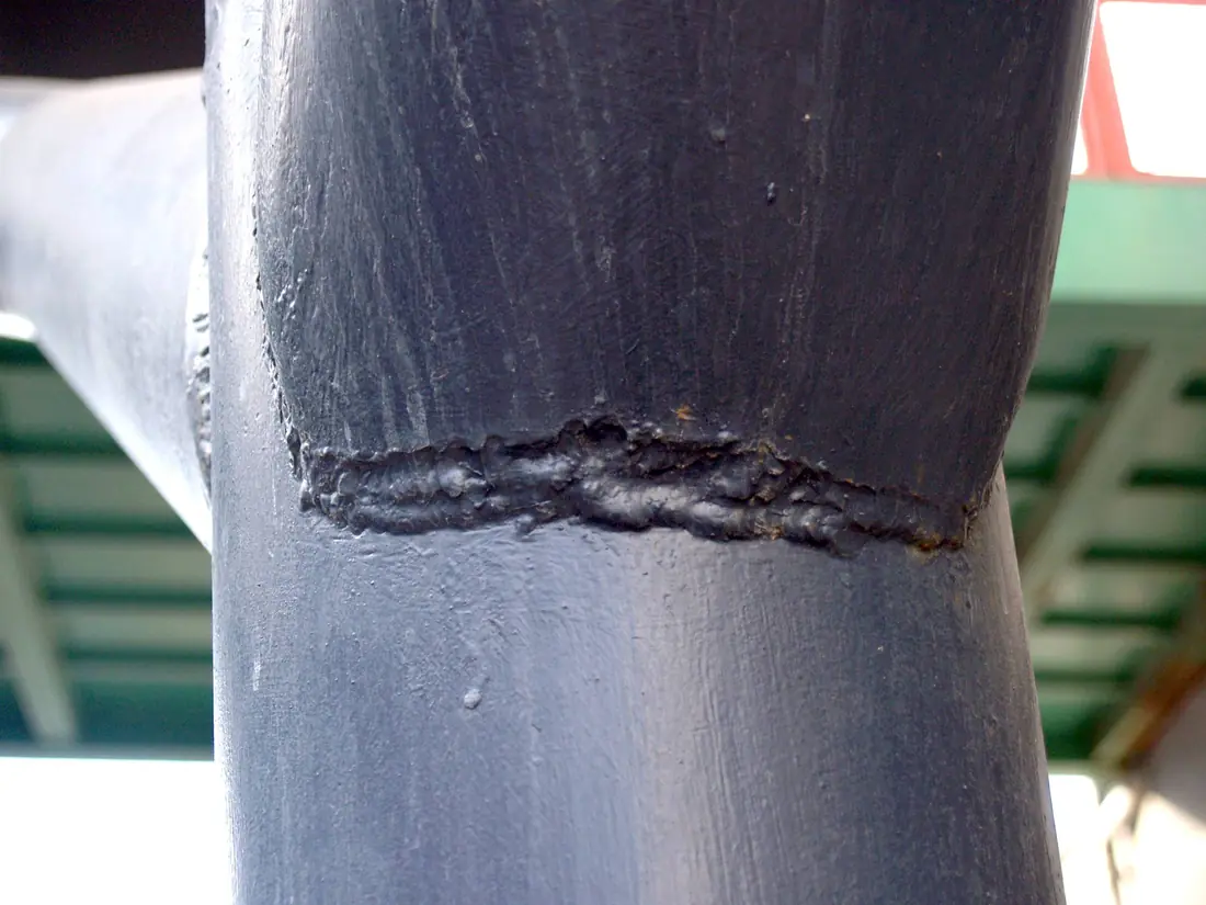 Painted steel pipe joint with a rough, lumpy weld bead of uneven width wrapped around it.
