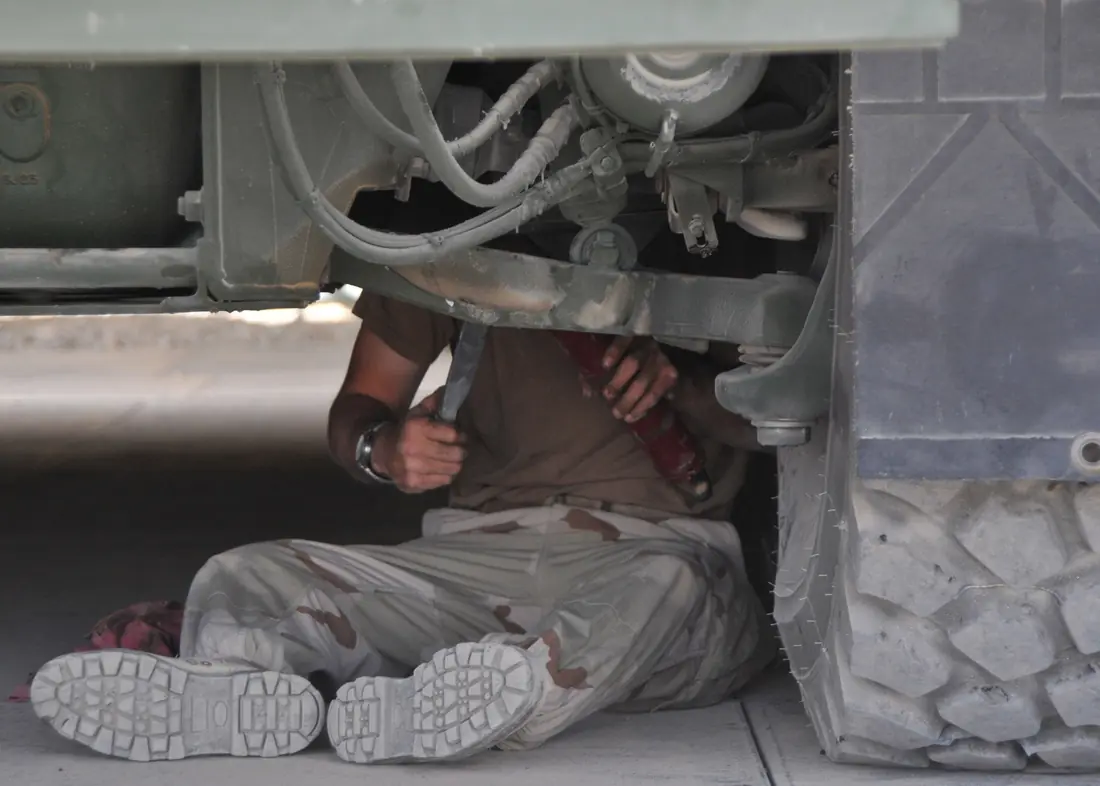 A mechanic sitting under a truck, working a grease gun onto a chassis fitting.