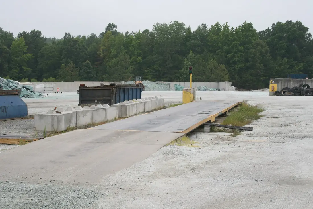 Empty in-ground truck scale platform at a materials yard, with concrete barriers and stockpiles behind it.