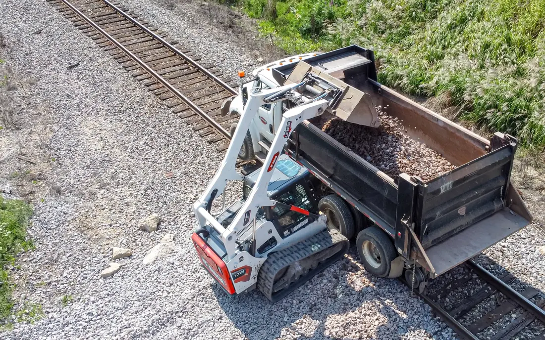 Overhead view of a compact track loader dumping a bucket of gravel over the side of a dump body.