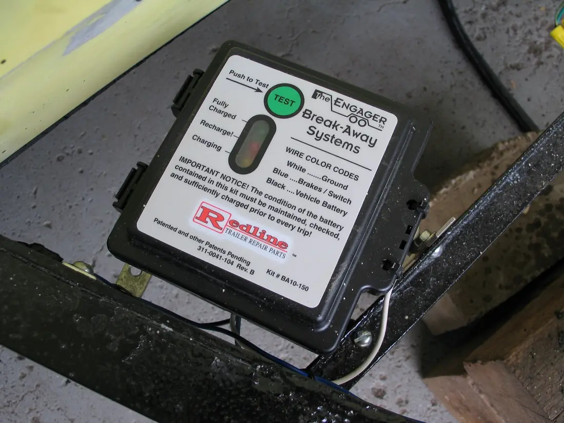 Black breakaway battery box bolted to a trailer frame, with a green push-to-test button and a charge indicator window.