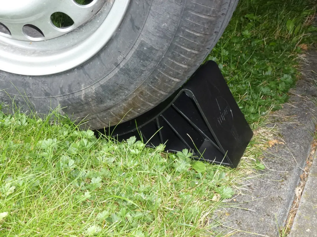 Black wheel chock wedged tight against the tread of a trailer tire on grass.